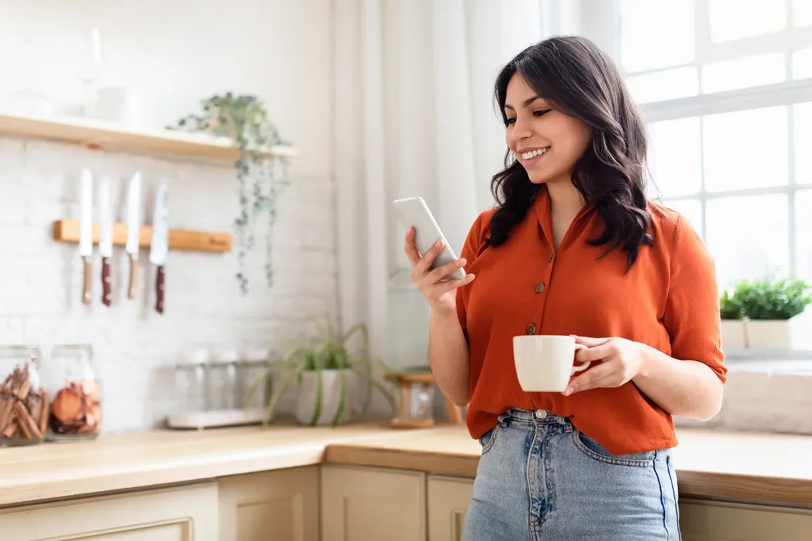 Caretaker in her kitchen, smiling at her phone with a coffee in hand