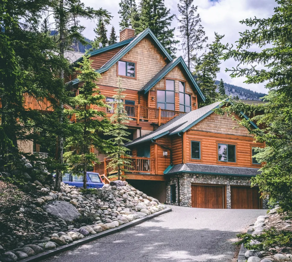Log cabin with green trim among pine trees, with a stone driveway and garage