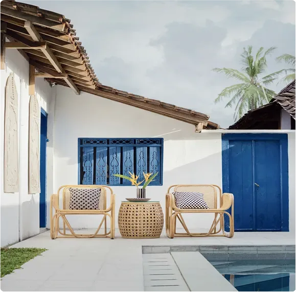 A shaded patio with rattan chairs outside a white house with blue doors