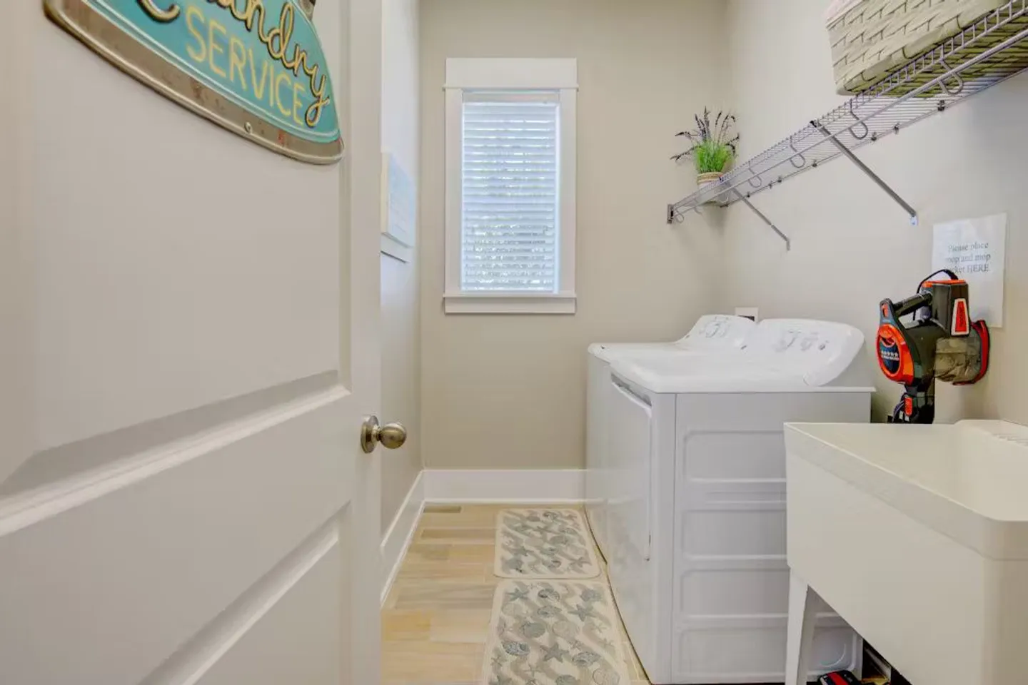 A laundry room viewed through an open doorway, featuring a white washer, dryer, utility sink, and a wire shelf above.