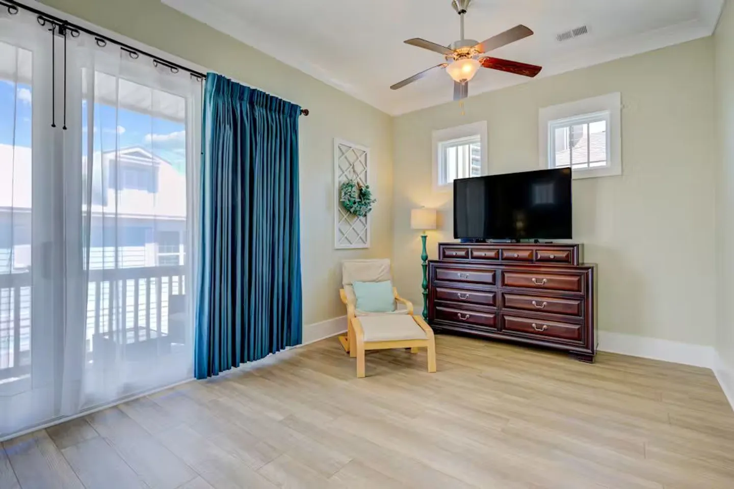 Bedroom or sitting area with wood-look floors, a ceiling fan, a large dark wood dresser with a TV, an armchair, and glass doors with teal curtains.