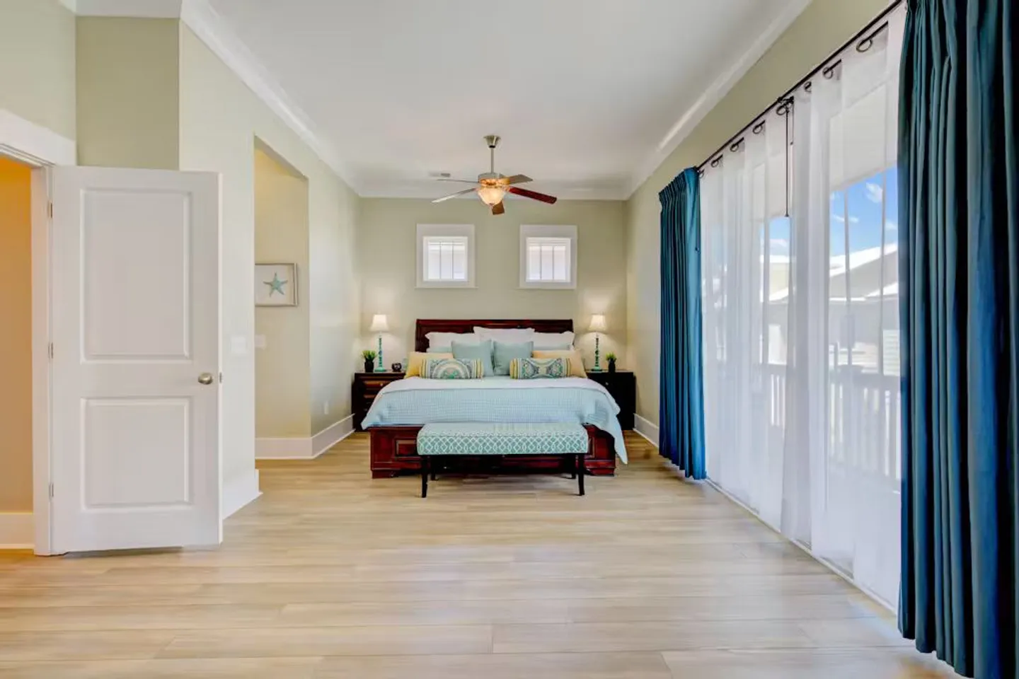 Spacious bedroom with a dark wood bed frame, light blue bedding, pale green walls, and large sliding doors covered with sheer and teal curtains.