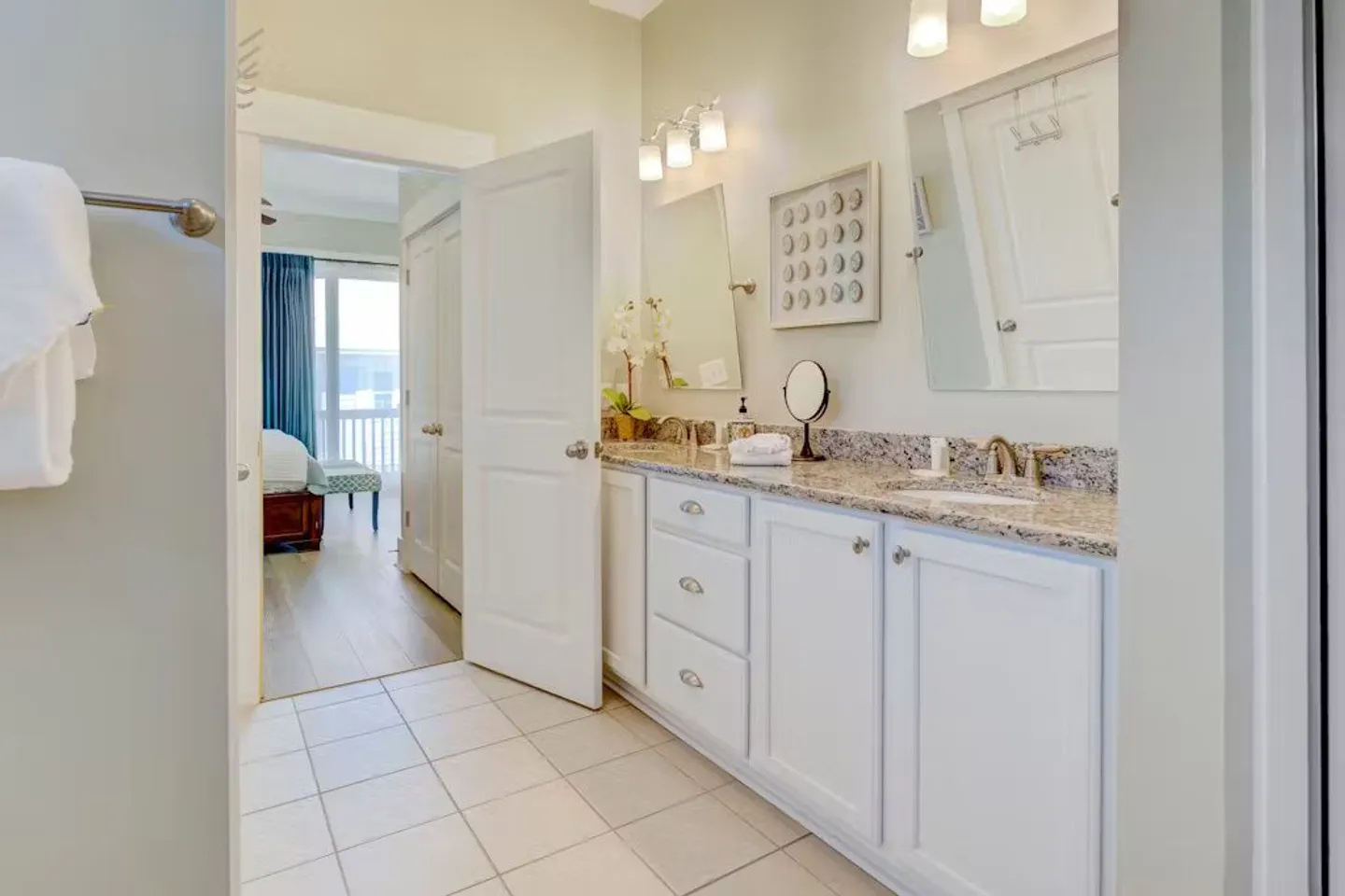 Bathroom with white double vanity, granite countertop, two mirrors, tile flooring, and doorway leading to an adjoining bedroom.