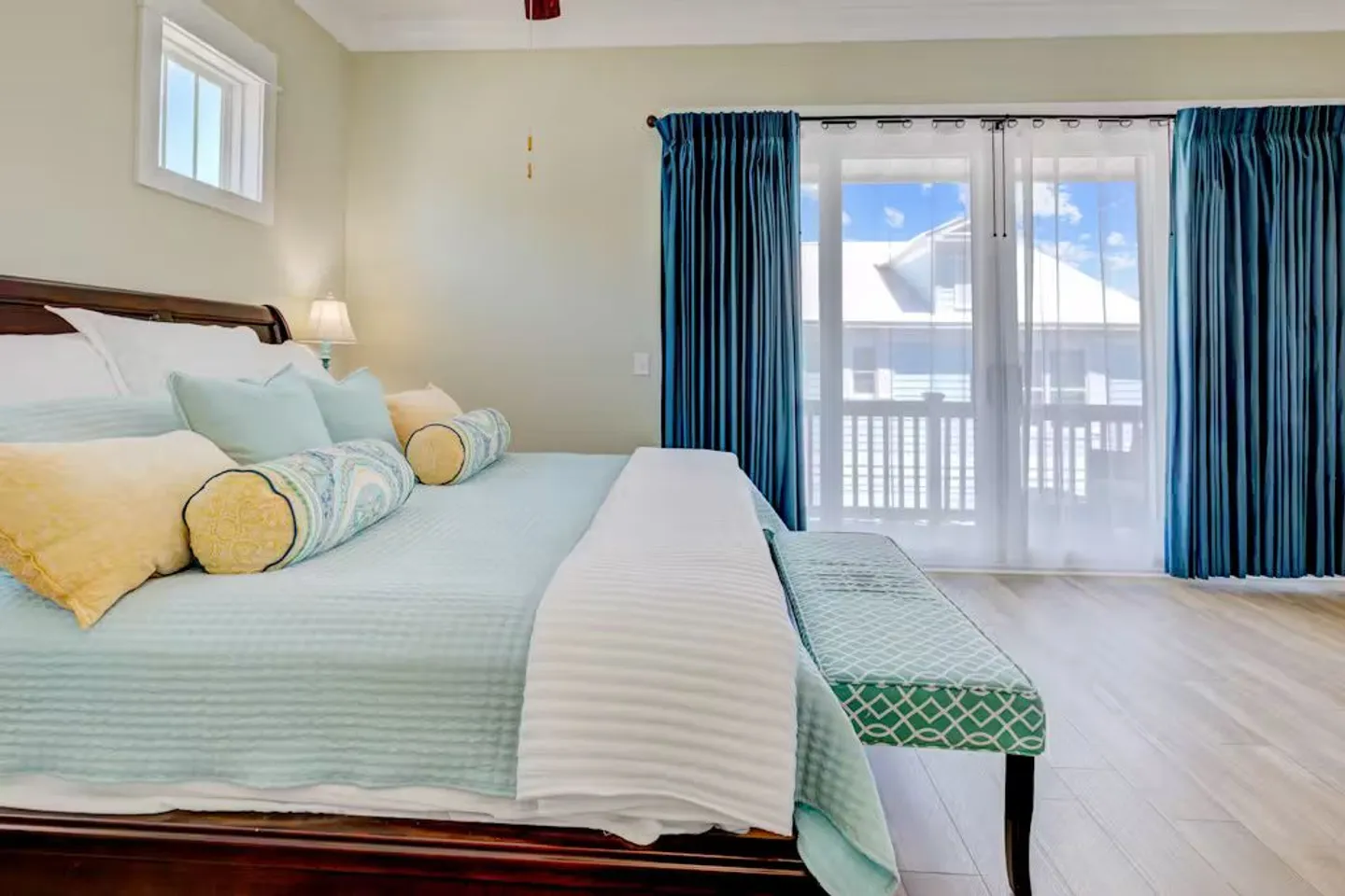 Bedroom with a large bed dressed in light blue and yellow pillows, an upholstered bench, and sliding glass doors leading to a balcony.