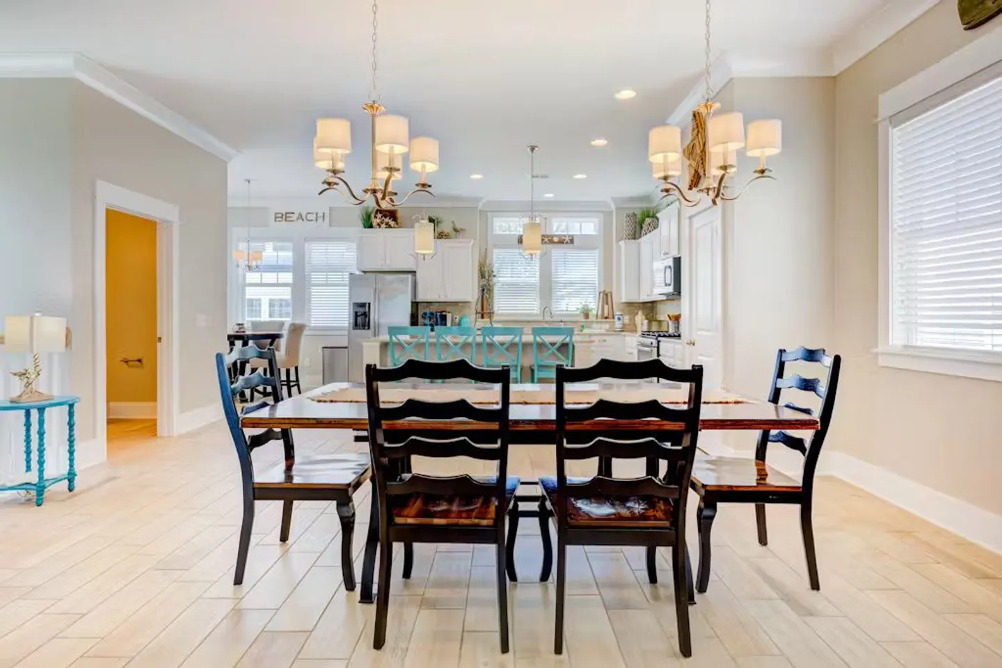 Open-concept dining area with a long wooden table and dark chairs, leading into a kitchen with an island and turquoise barstools.