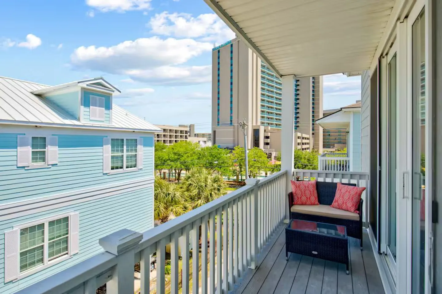 Covered outdoor balcony with a bench, coffee table, and railings overlooking neighboring homes and a multistory building under a blue sky.