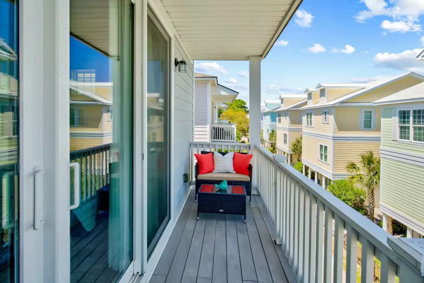 Covered outdoor balcony with a bench, coffee table, and views of neighboring pastel-colored homes under a bright blue sky.