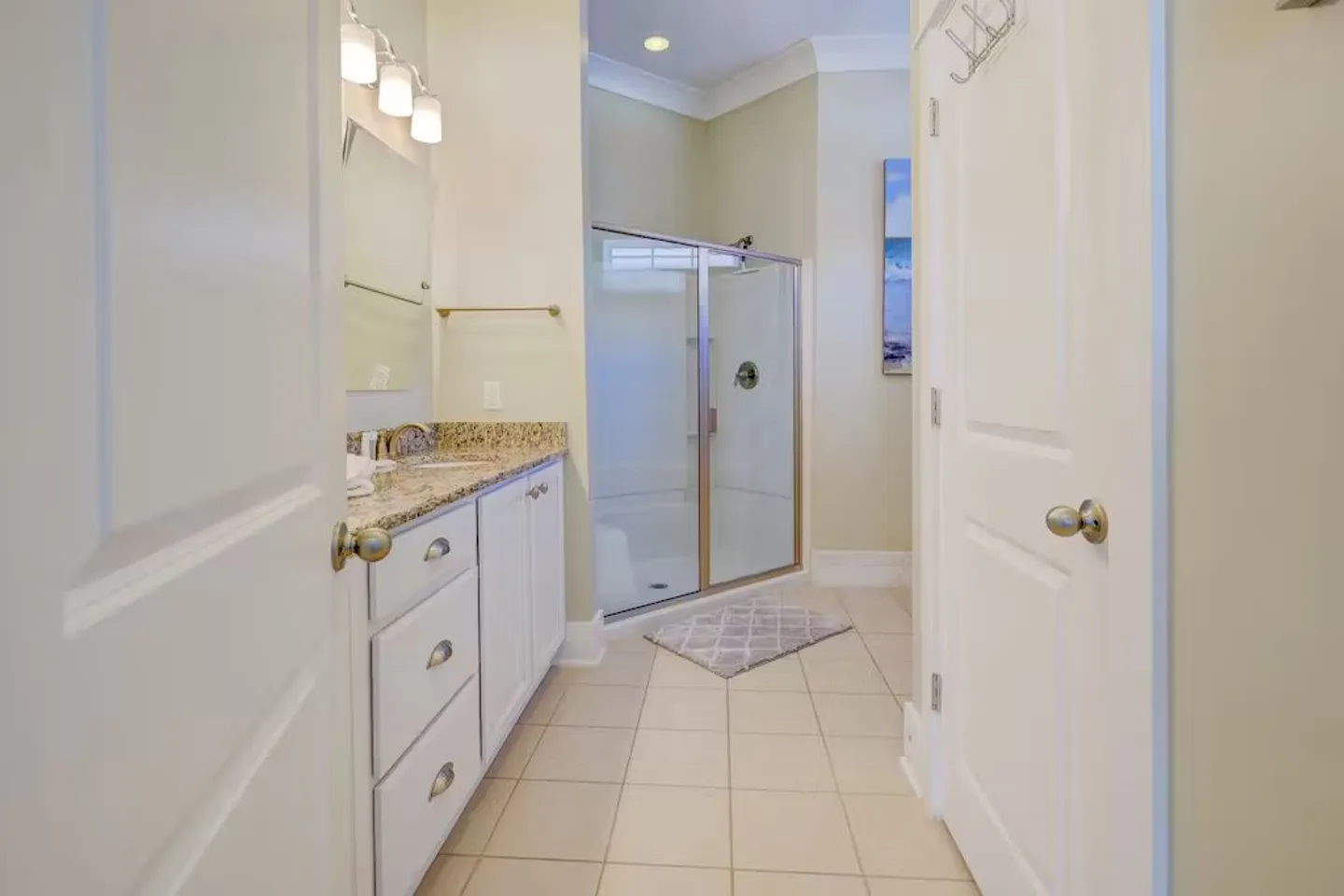 Bathroom viewed through an open doorway, featuring a vanity with speckled countertop and white cabinets, tiled floor, and a walk-in shower with sliding glass doors.