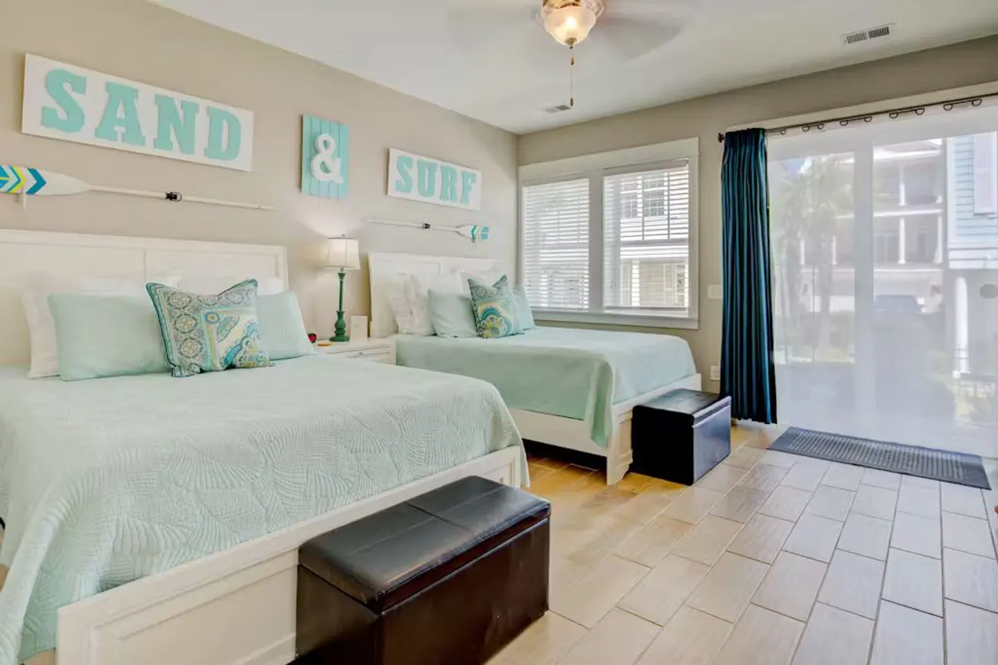 Bedroom featuring two beds with light turquoise bedding, dark benches at the foot of each, tiled flooring, and sliding glass doors to the outside.