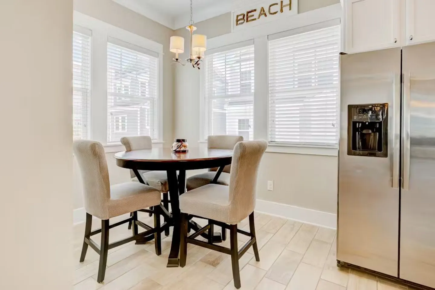 Dining nook with a round table and upholstered chairs beside windows with horizontal blinds next to a stainless steel refrigerator.