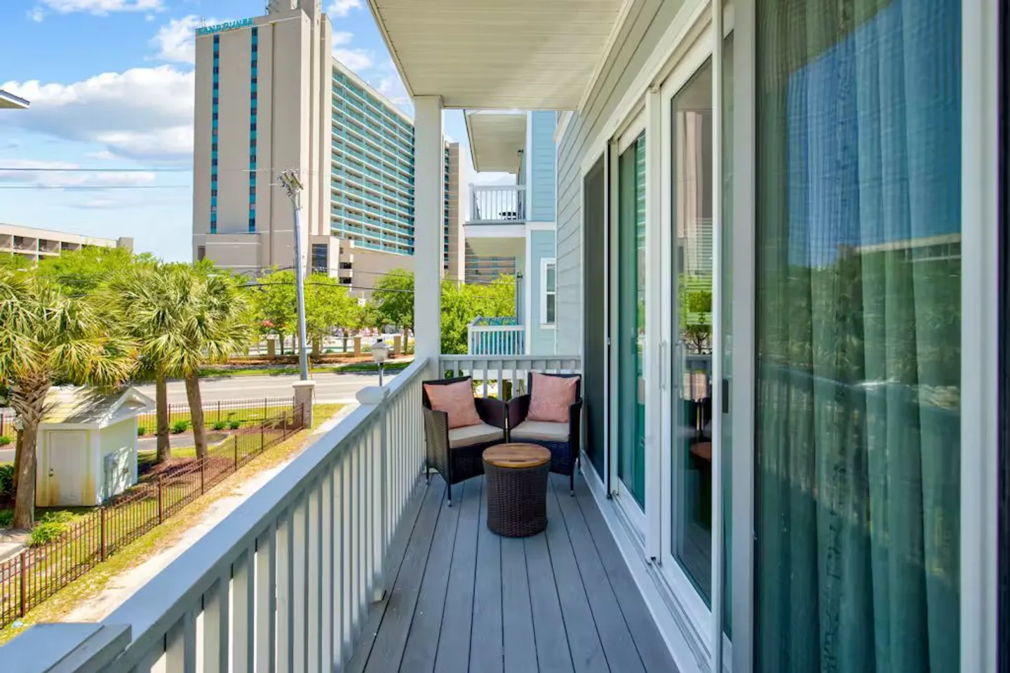 Covered outdoor balcony with wood decking, railing, two wicker armchairs, and a small round table facing surrounding buildings.