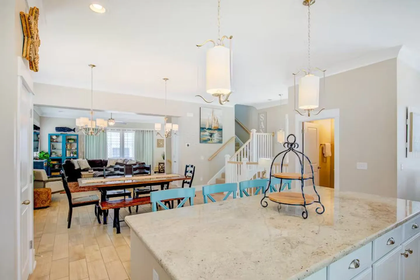 Open-concept interior featuring a kitchen island with light blue stools in the foreground, leading to a dining table and living room in the background.