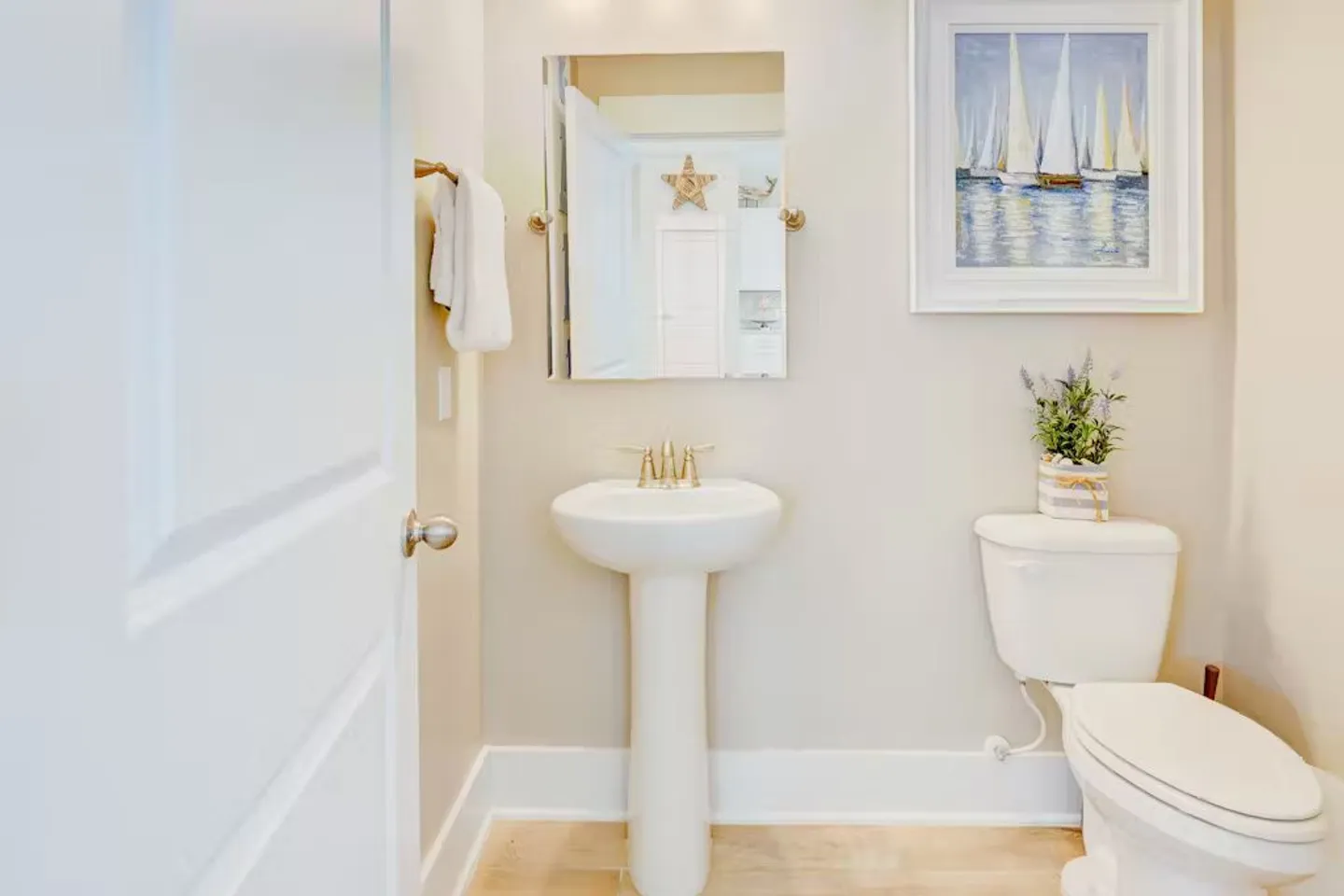 Powder room featuring a white pedestal sink with brass fixtures, a mirror, and a white toilet beneath framed sailboat artwork.