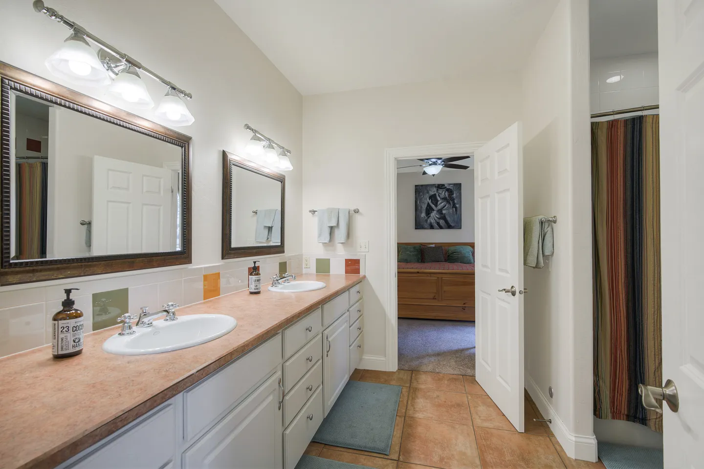 Bathroom with double vanity, two framed mirrors, overhead lighting, and an open doorway leading to a bedroom.