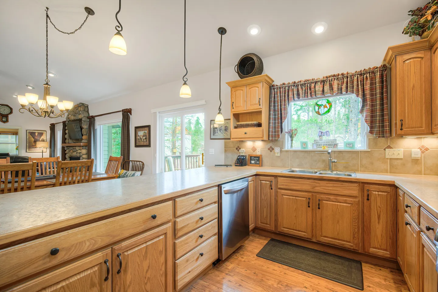 Kitchen with wooden cabinets, laminate countertops, and stainless steel dishwasher looking toward dining area and stone fireplace.