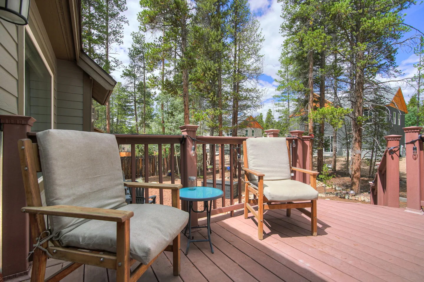 Wooden deck with two cushioned armchairs and a small side table overlooking tall pine trees under a blue sky.