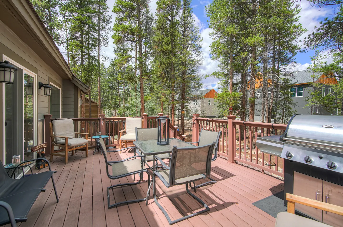 Spacious outdoor wooden deck with a patio dining table, chairs, a barbecue grill, and surrounding pine trees.