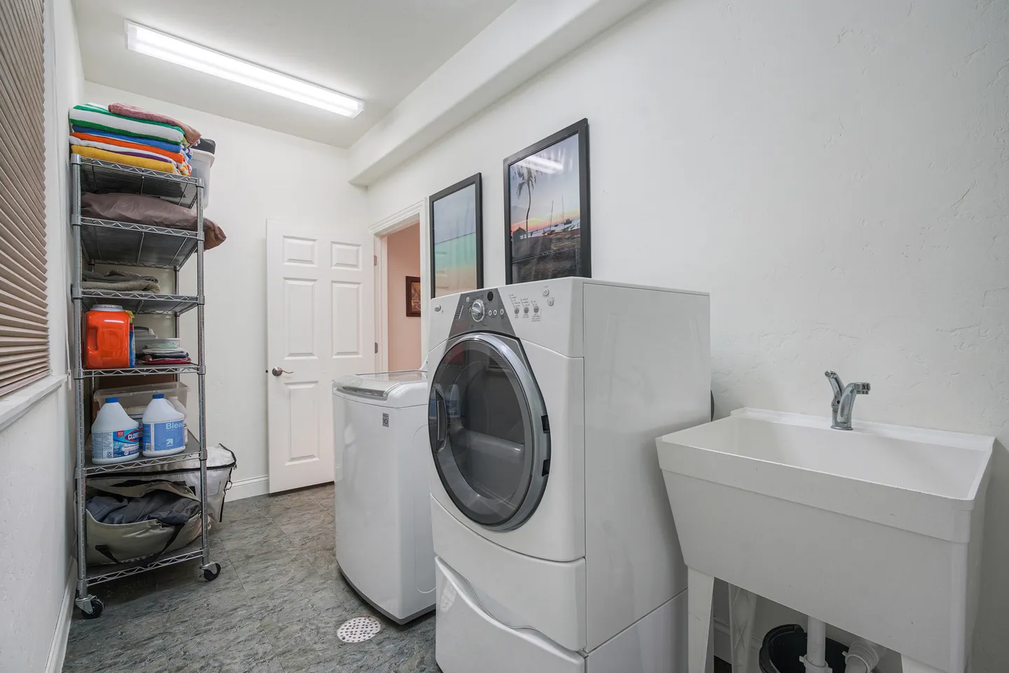 A brightly lit laundry room with a white washing machine, dryer, utility sink, and metal storage rack.