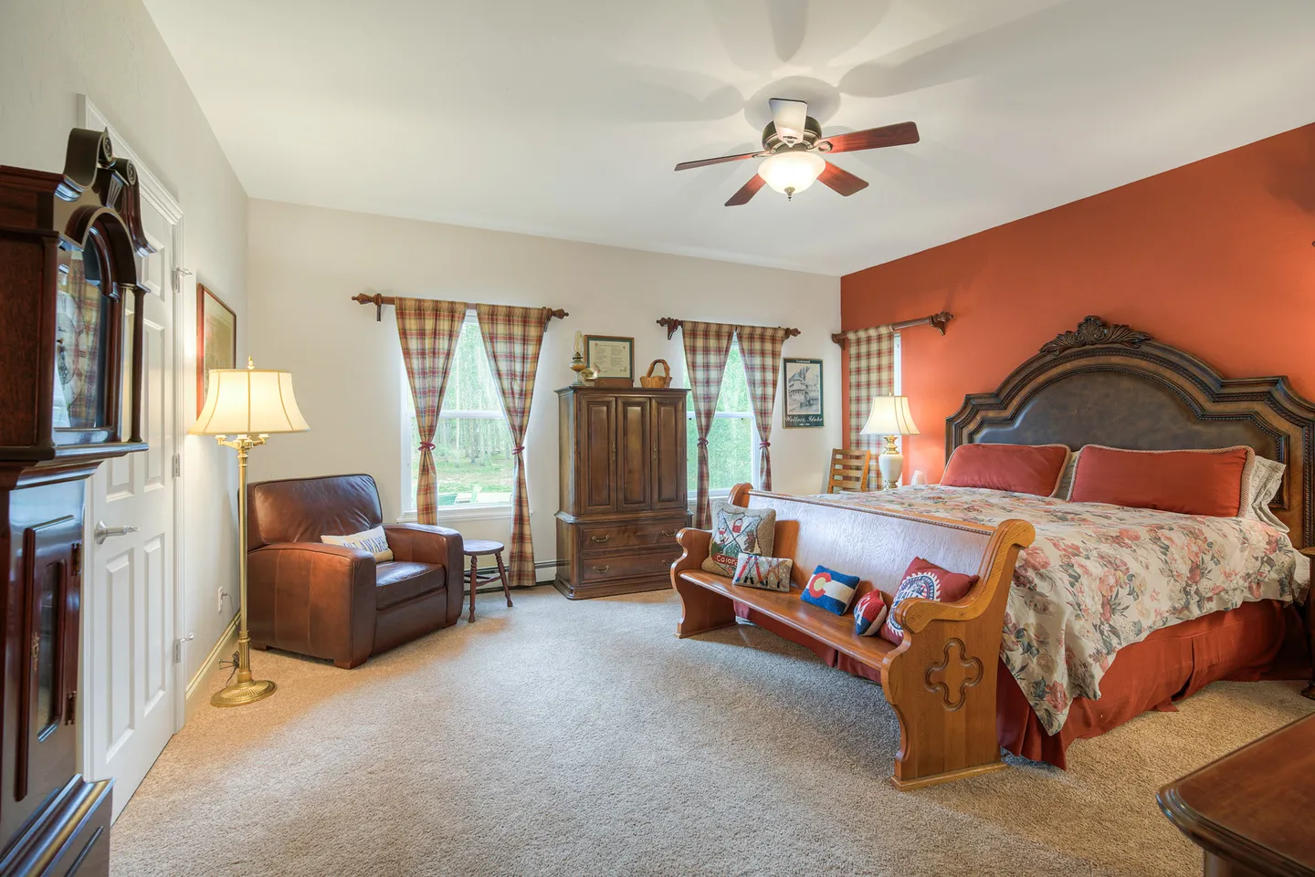 Bedroom with carpet, an ornate bed against a terracotta accent wall, a wooden bench, leather armchair, grandfather clock, and wardrobe.