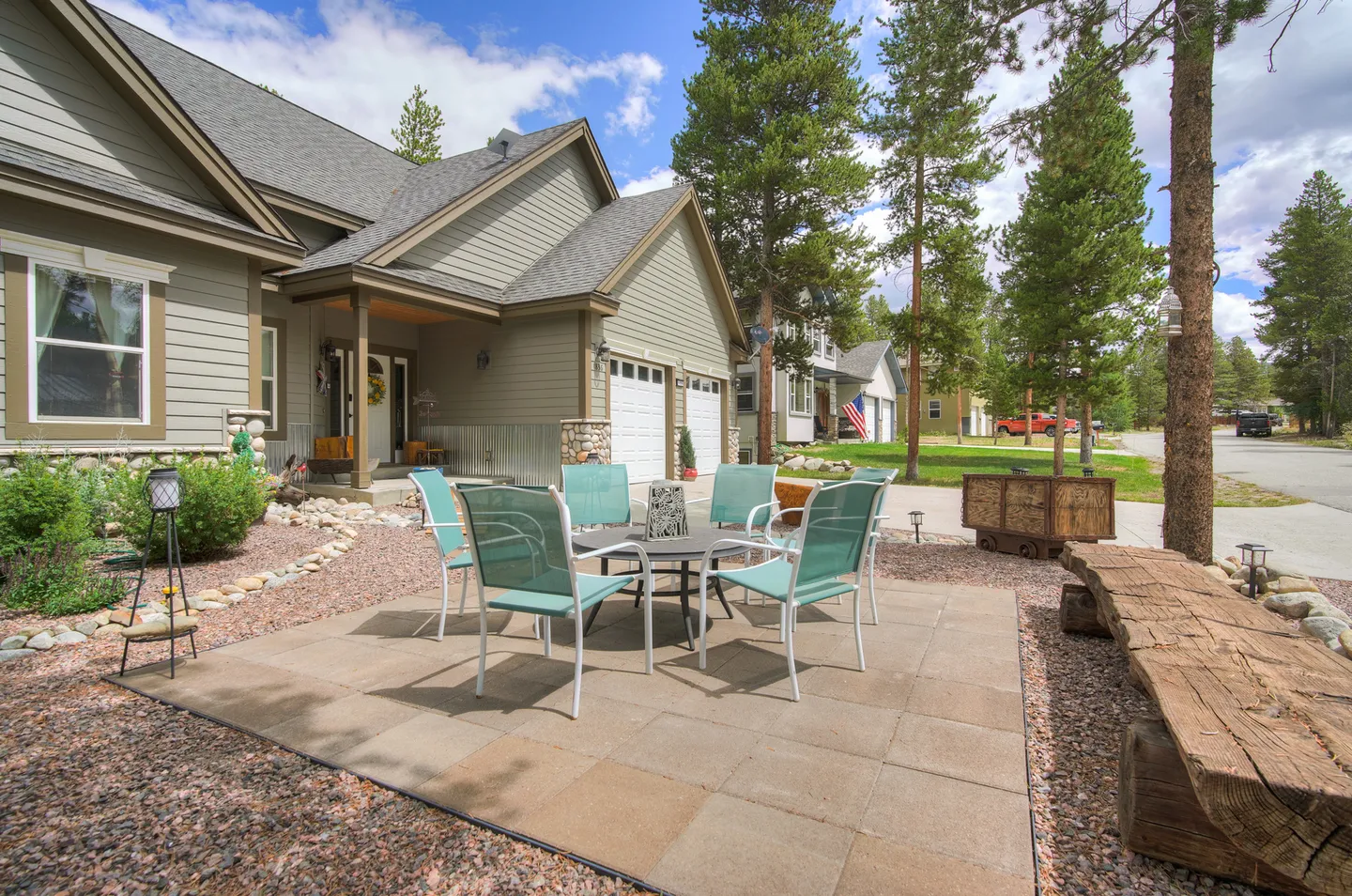 Outdoor patio with a table and chairs in front of a multi-level house bordered by pine trees.