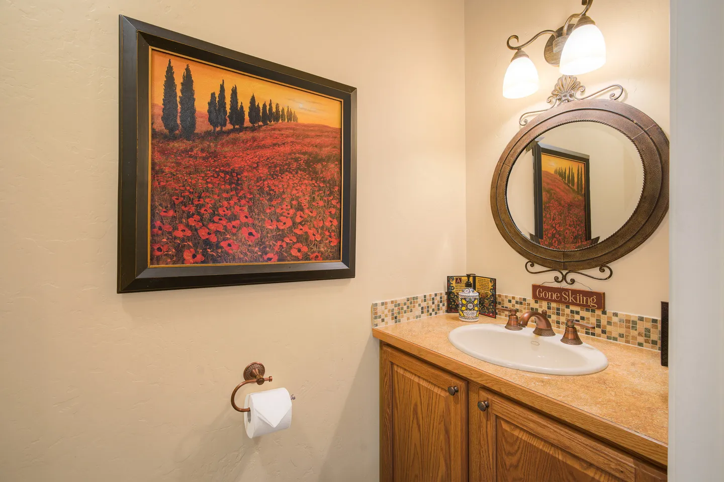 Powder room featuring a wooden vanity, oval mirror, framed poppy field artwork, and warm wall lighting.