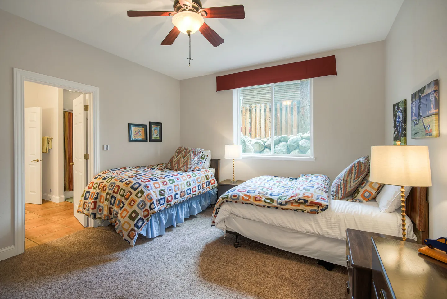 Bedroom with two twin-style beds, patterned colorful bedding, carpeted floor, ceiling fan, and window looking out to a retaining wall.