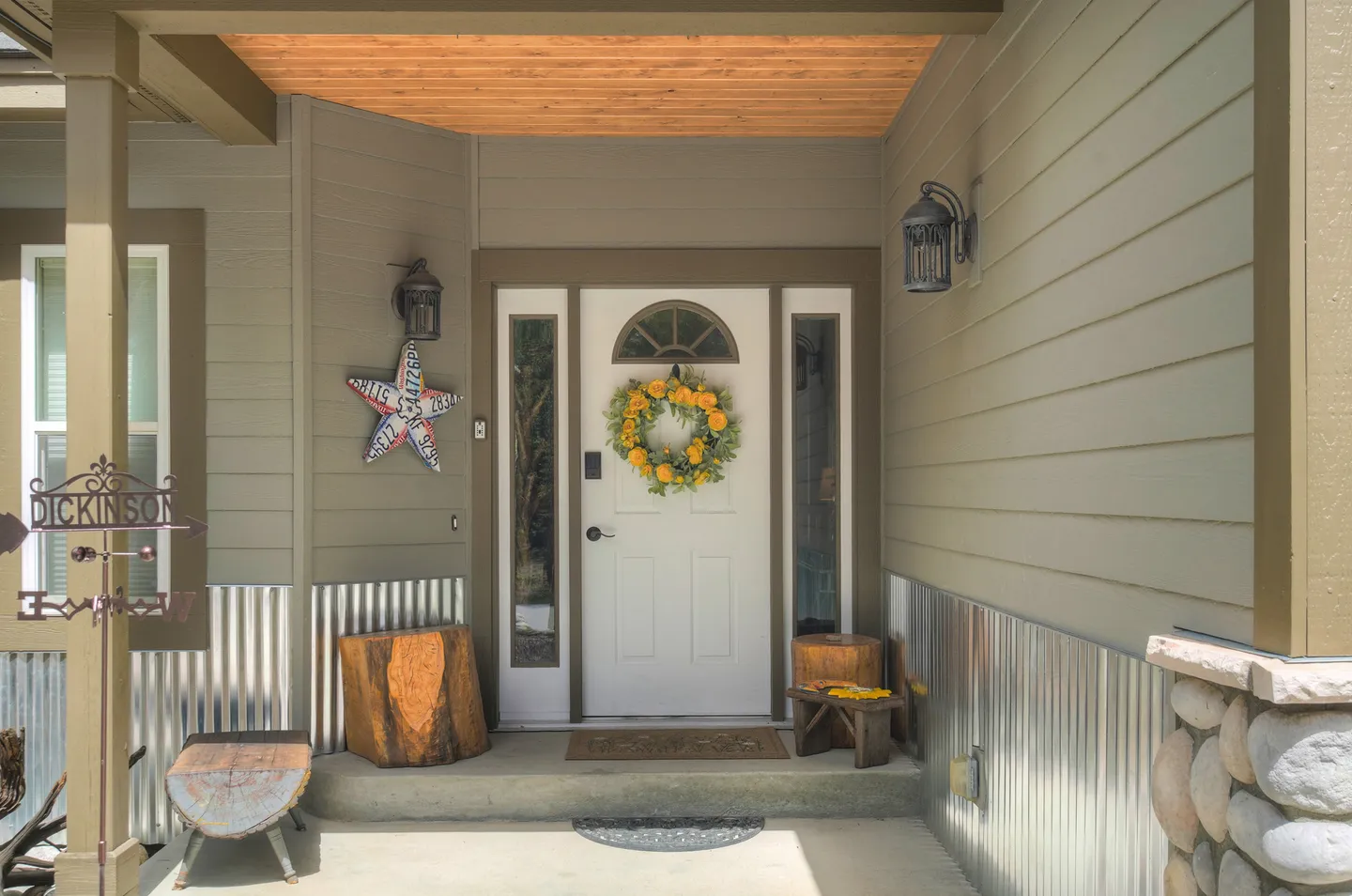 Front porch and entrance of a home featuring a white front door adorned with a yellow wreath, corrugated metal wainscoting, and rustic accents.