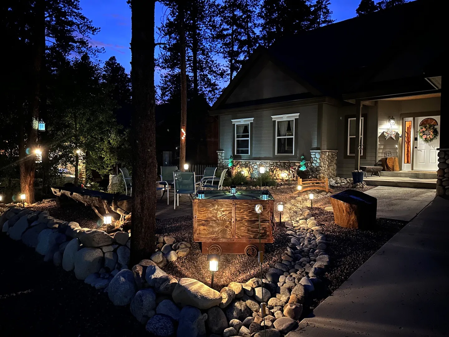 Nighttime view of a house exterior and front yard illuminated by landscape lighting with rock-lined borders and a seating area.