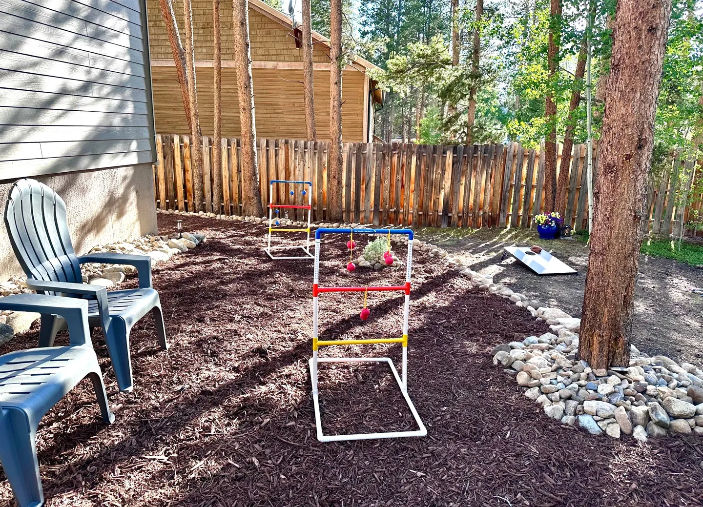 Outdoor mulched play area with a ladder toss game, cornhole board, and plastic chairs alongside a wooden fence.