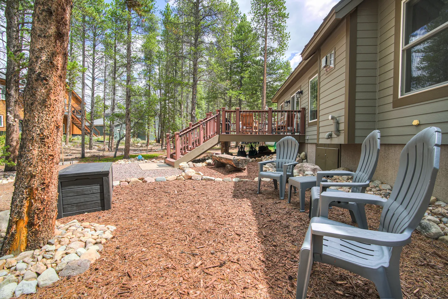 Mulched yard beside a house with gray Adirondack chairs, an elevated wooden deck, and tall pine trees.