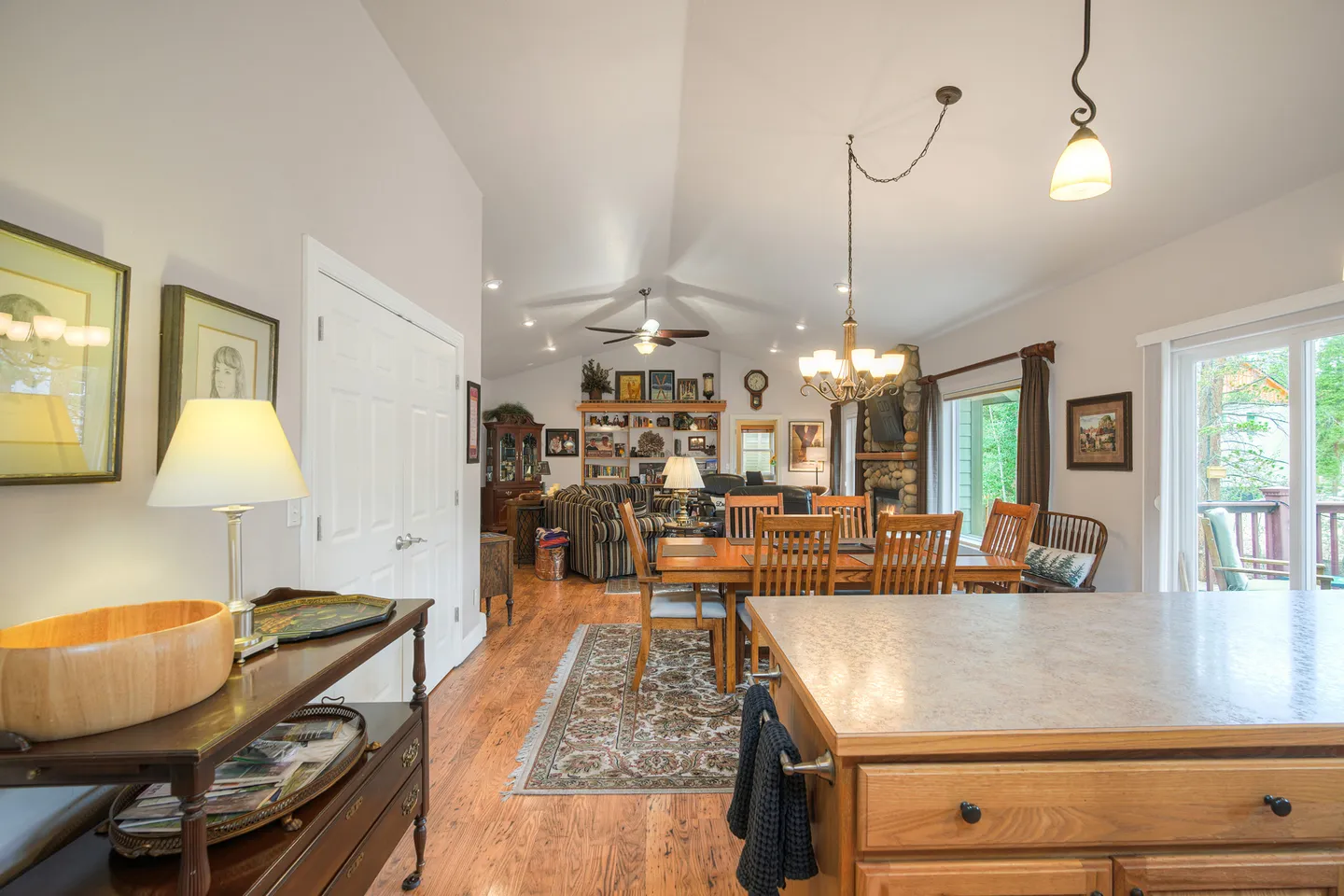 Open-concept living and dining area viewed from the kitchen, featuring vaulted ceilings, wood floors, a dining table with chairs, and a living room in the background.