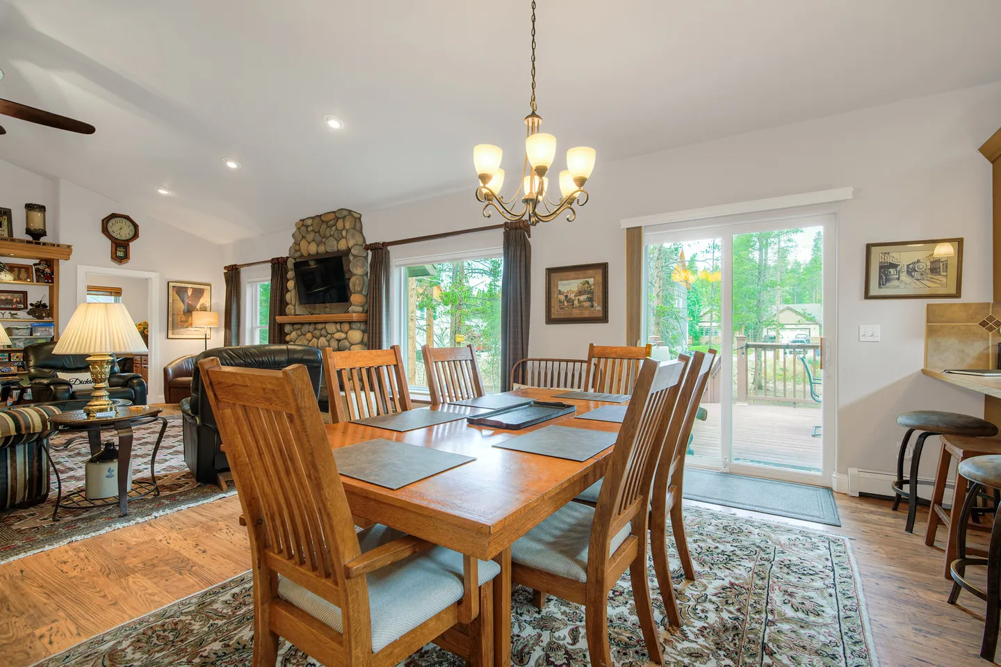 Dining area with a wooden table and chairs on a patterned rug, adjacent to a living area with a stone fireplace and sliding glass doors to a deck.
