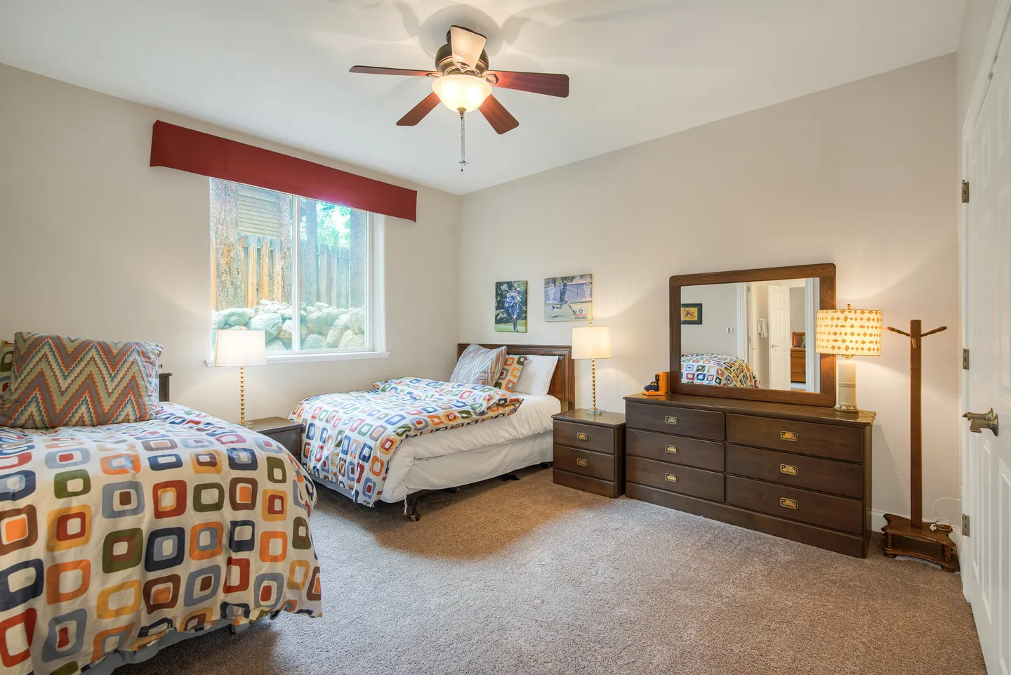 Bedroom featuring two twin beds with colorful geometric bedding, carpeted floor, ceiling fan, and a wooden dresser with a mirror.