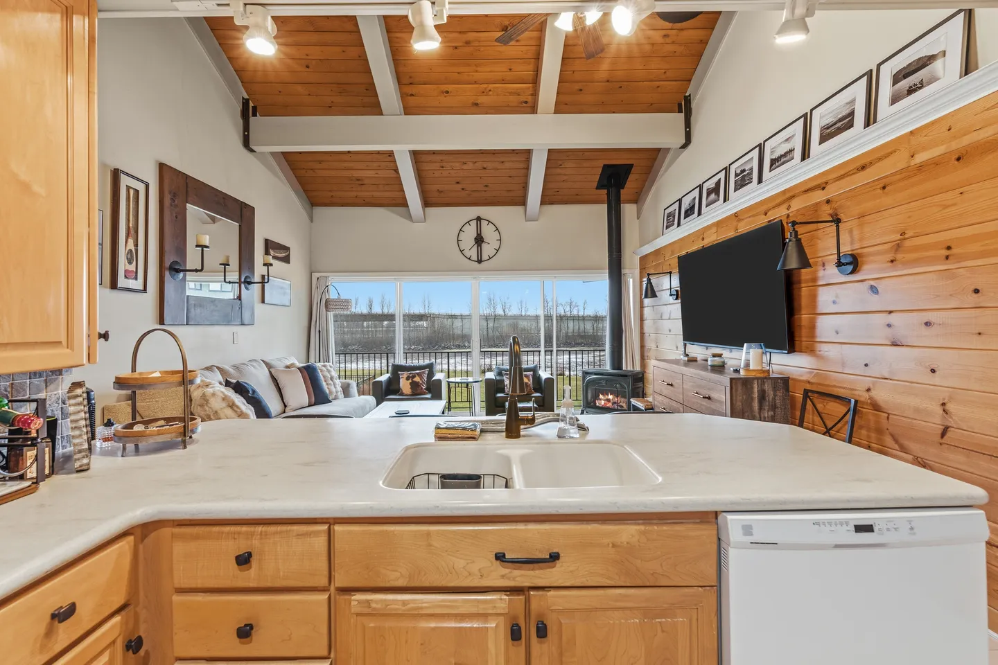View over a kitchen counter with a sink toward an open living room with vaulted wood ceilings, a fireplace, and large sliding glass doors.