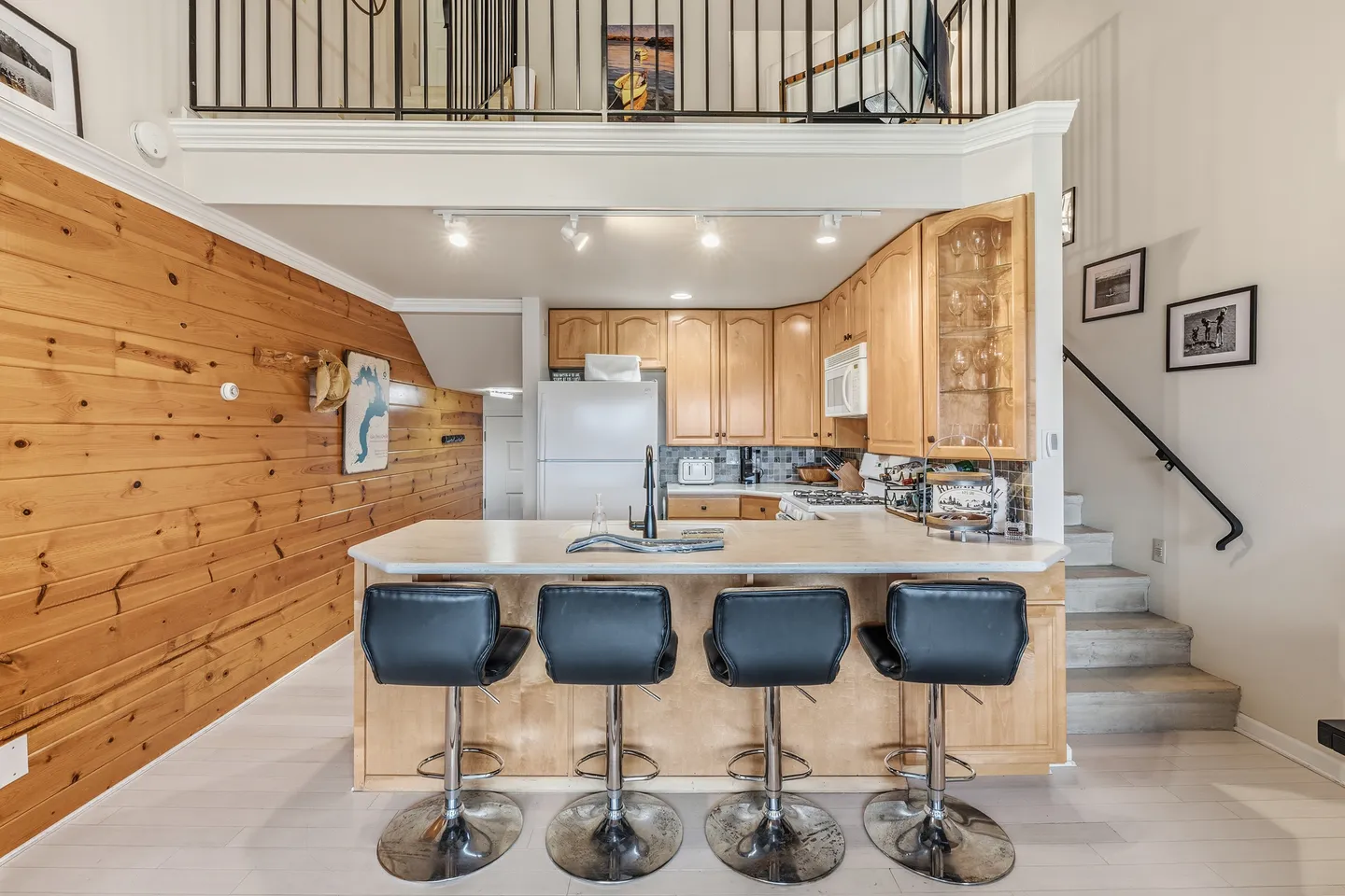 Kitchen with breakfast bar and four black stools, wooden paneled accent wall, and an open loft area above.