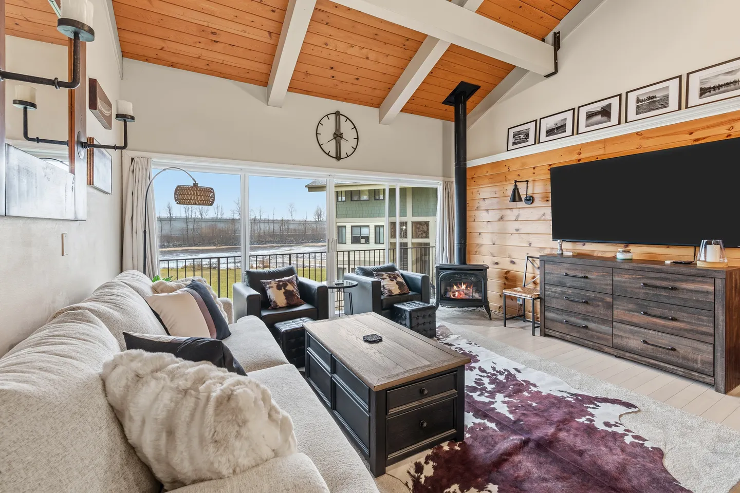 Living room with vaulted wood-paneled ceiling, comfortable seating, fireplace, large wall-mounted TV, and sliding glass doors to a balcony.