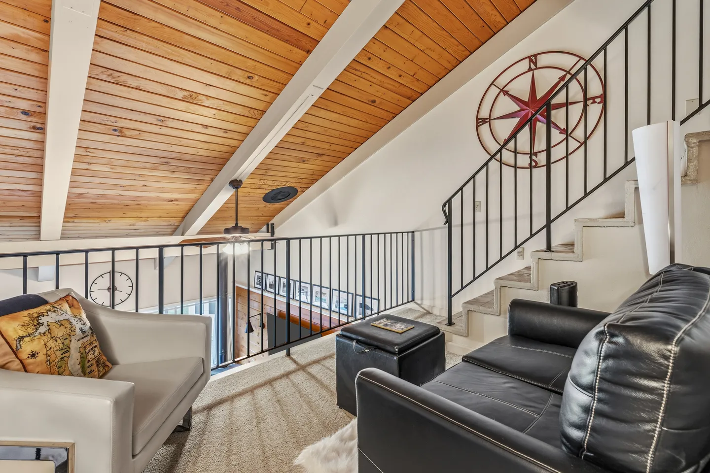 Upstairs loft sitting area with a black leather loveseat, beige armchair, wood-paneled vaulted ceiling, and stairs leading up.