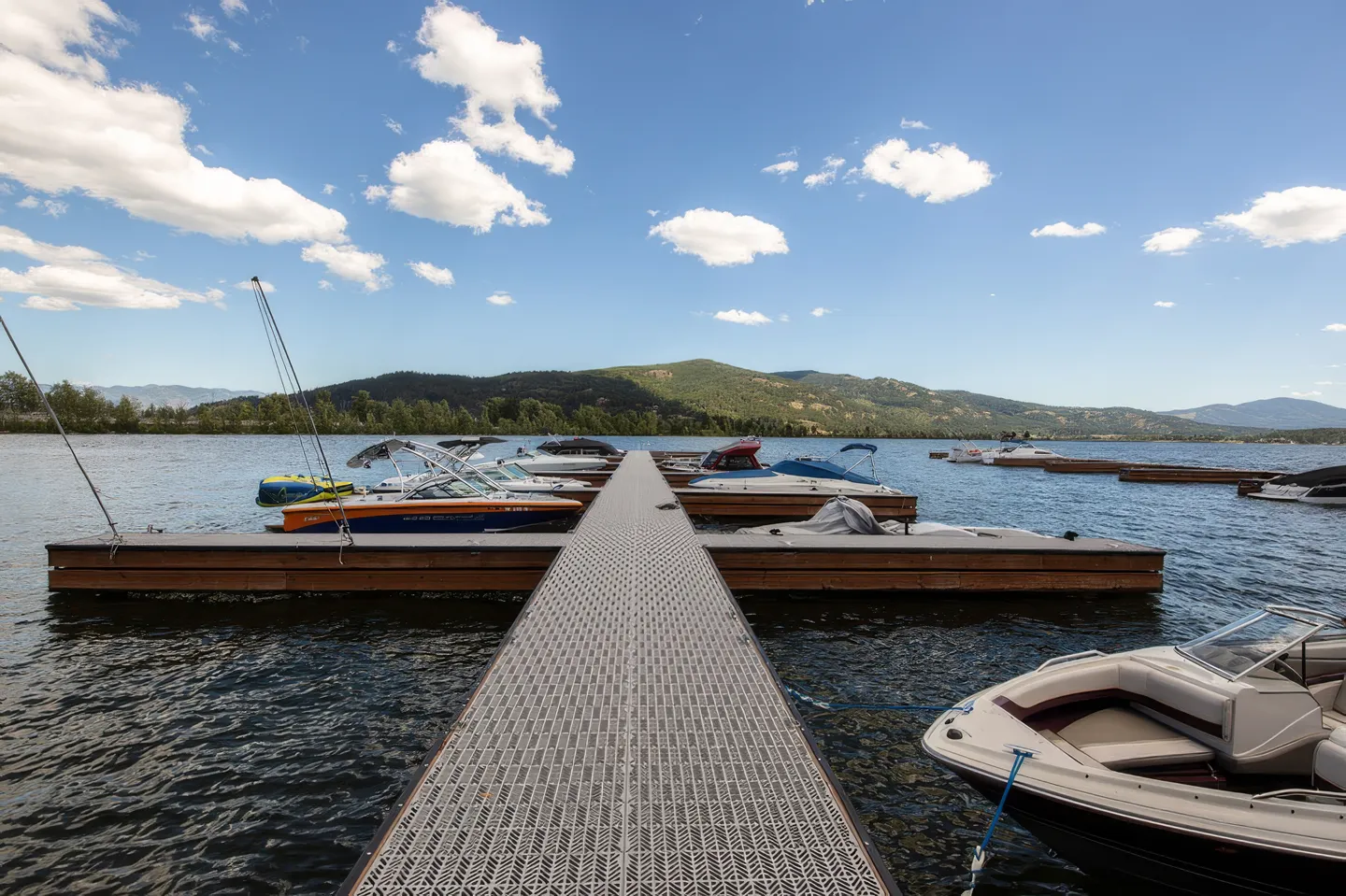 A floating boat dock on a lake with several moored boats and mountains under a blue sky.