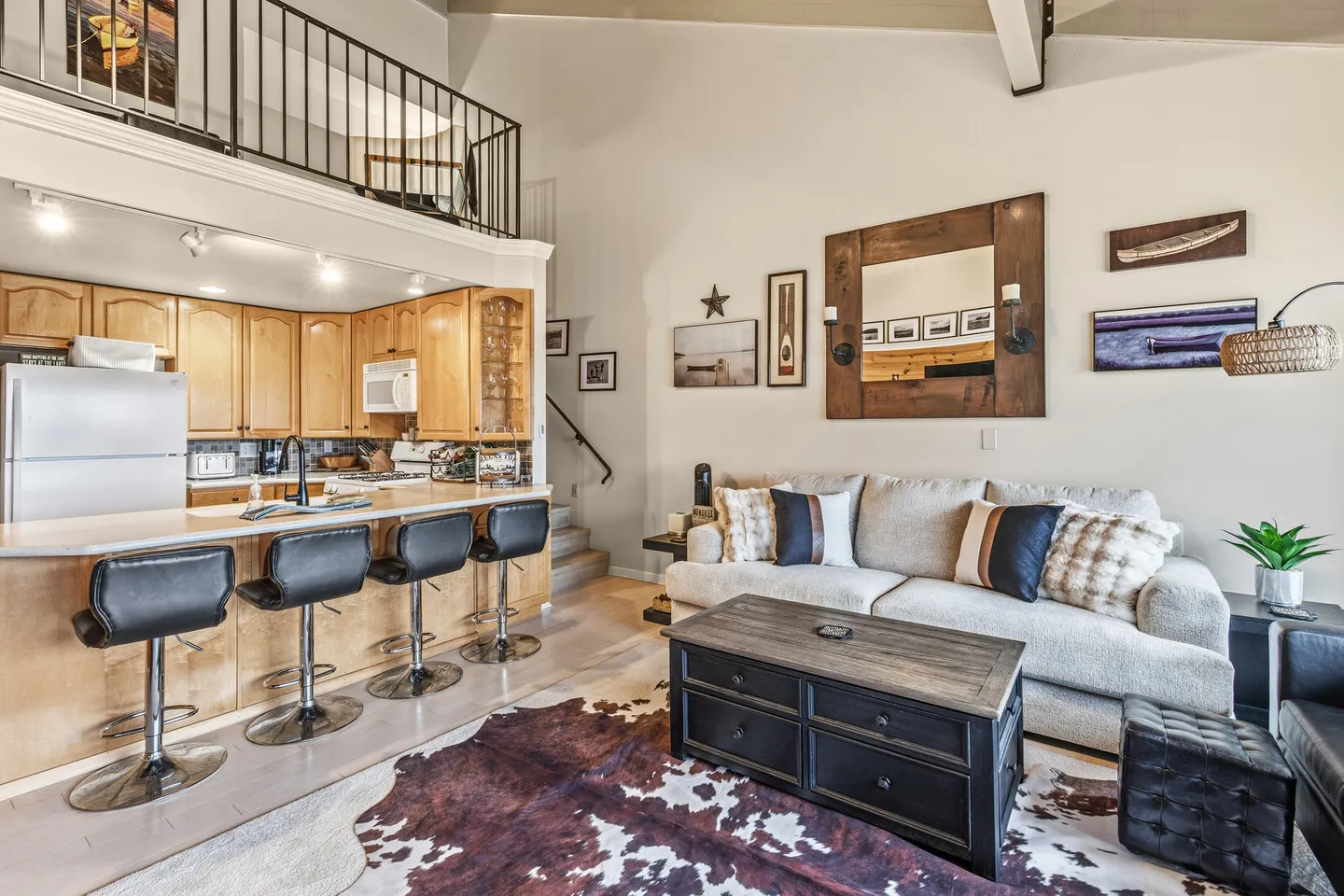 Open living area with a sofa, dark wood coffee table, cowhide rug, breakfast bar with barstools, and kitchen beneath an upper loft railing.