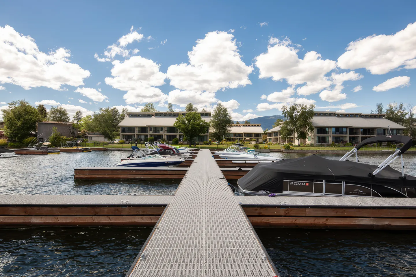 A floating boat dock extends across the water toward waterfront residential buildings under a blue, cloudy sky.