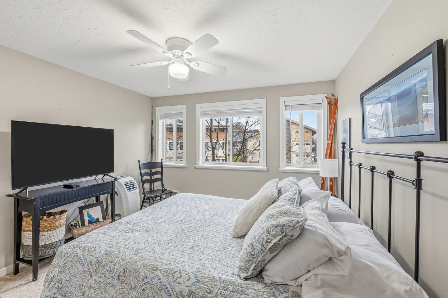 Bedroom featuring a made bed with an iron headboard, a ceiling fan, a flat-screen TV on a dark console table, and three windows overlooking trees and buildings.