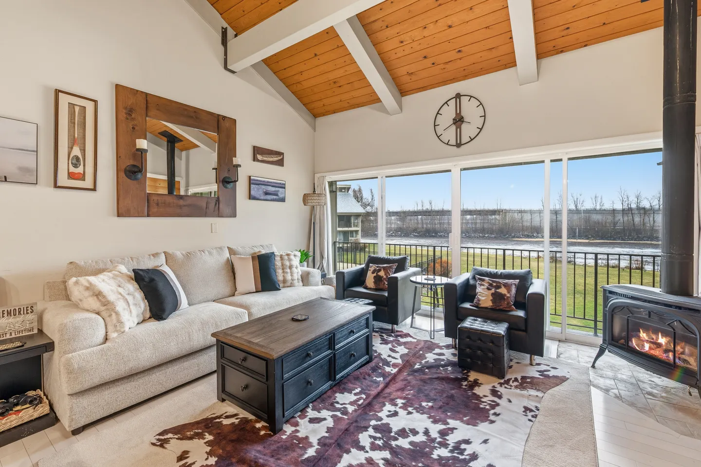 Living room with a sofa, two armchairs, coffee table, wood stove fireplace, vaulted wood-paneled ceiling, and sliding glass doors.