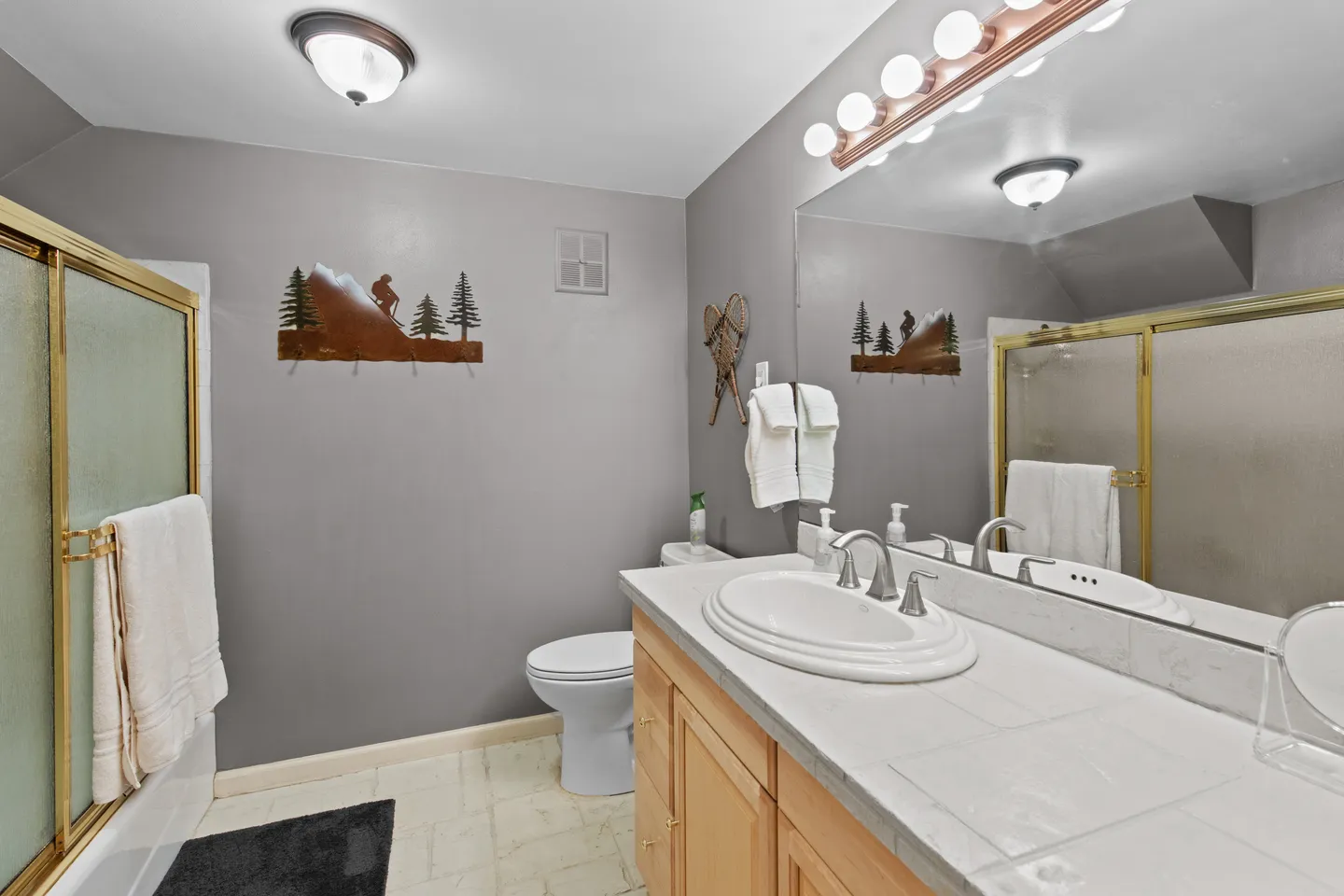 Bathroom with a wood vanity, white tile countertop, large mirror, toilet, and glass shower enclosure with brass-toned frame.