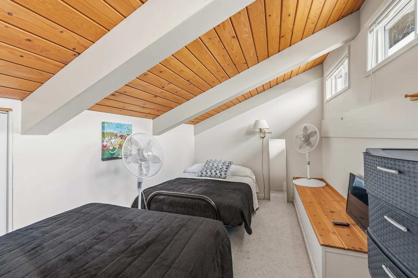 Loft-style bedroom with sloped wood-plank ceiling, exposed white beams, two beds, and two standing fans.