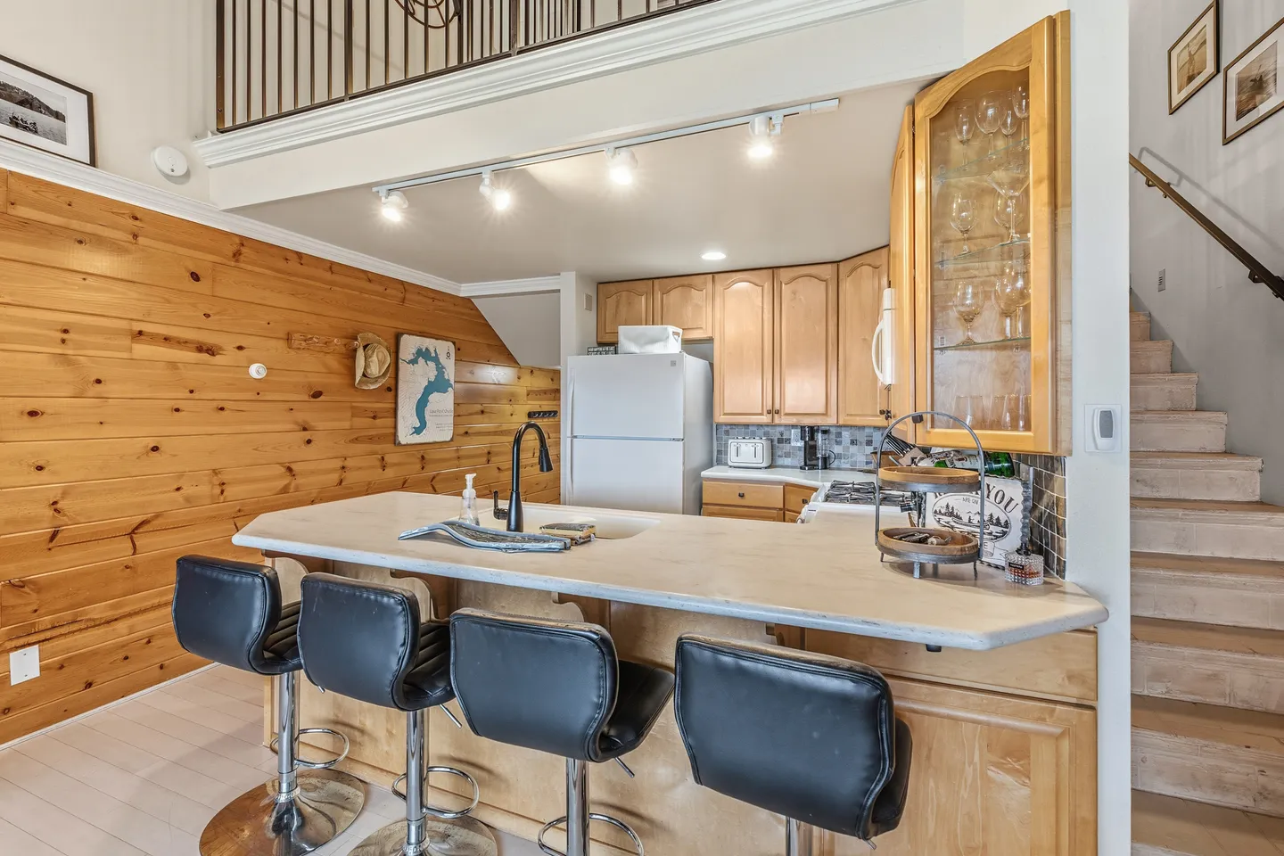 Kitchen with wooden cabinetry, breakfast bar with black stools, and a wood-paneled accent wall next to a staircase.