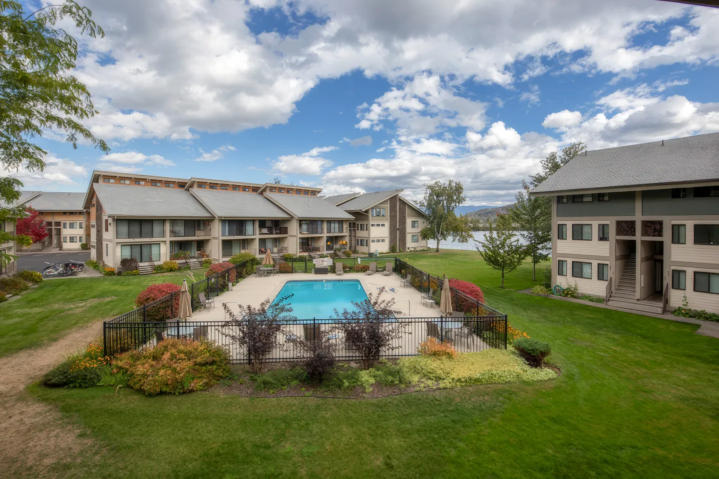 Elevated view of a fenced community swimming pool surrounded by lawn, landscaping, and multi-story condominium buildings near a lake.
