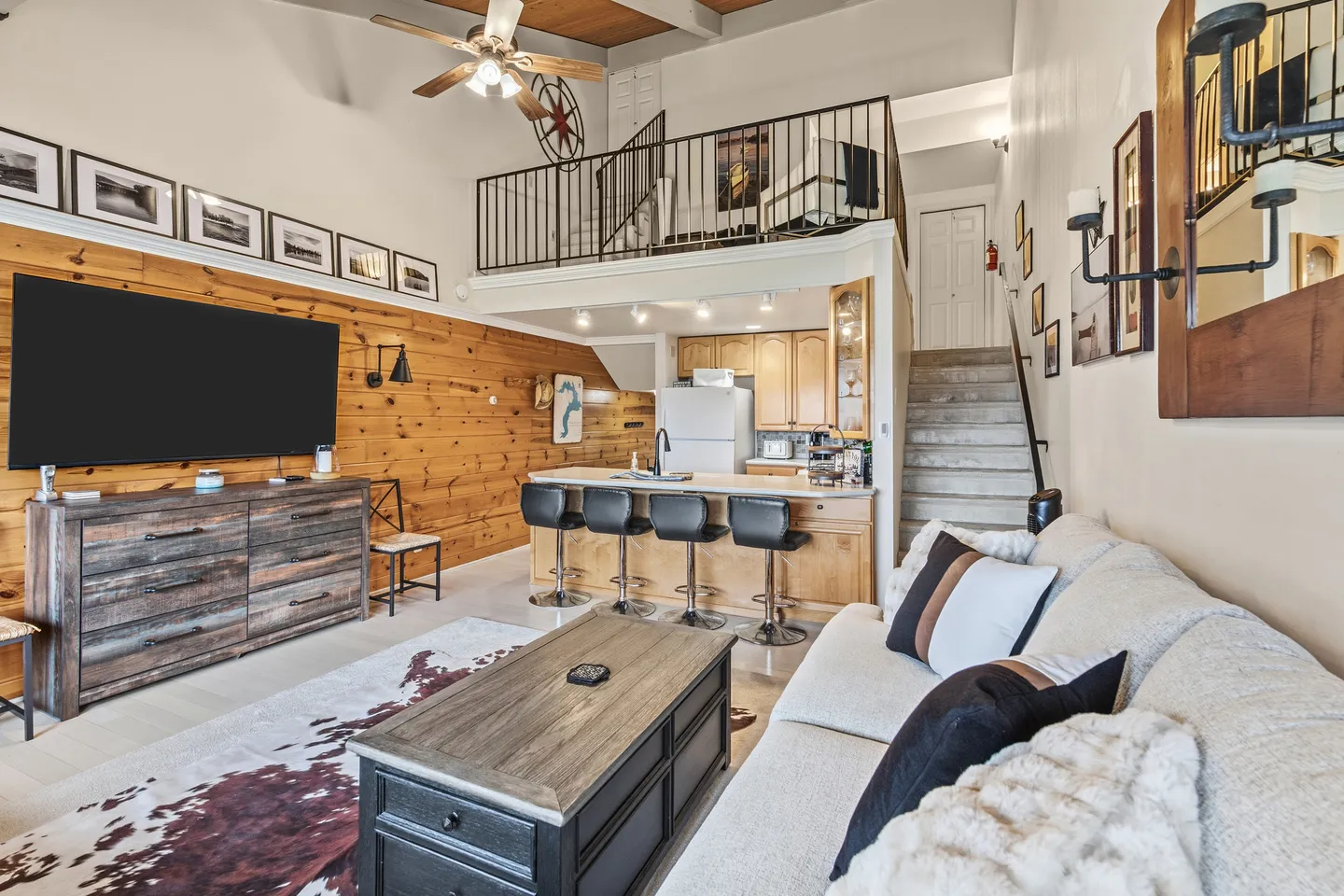 Open-concept living room with wood-paneled feature wall and TV, adjacent to a kitchen with breakfast bar below a loft.