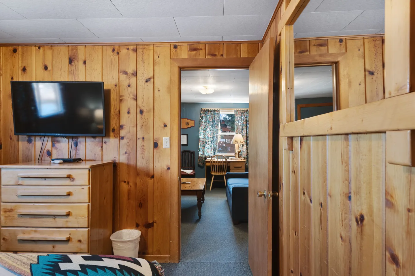 View from a wood-paneled bedroom with a dresser and mounted television looking into an adjoining carpeted living area.