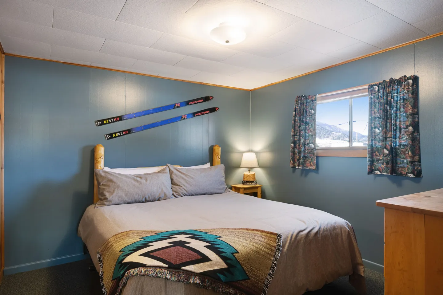Bedroom with blue walls, a bed flanked by a rustic headboard, decorative skis mounted on the wall, and a window.