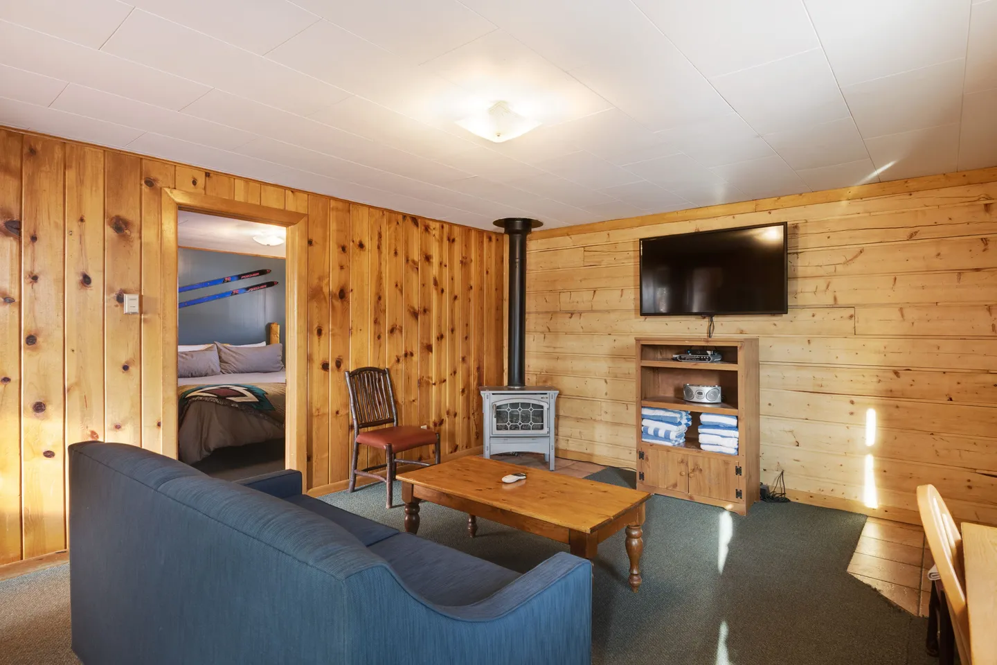Living room with wood-paneled walls, blue couch, coffee table, wood-burning stove, wall-mounted television, and a doorway opening to a bedroom.